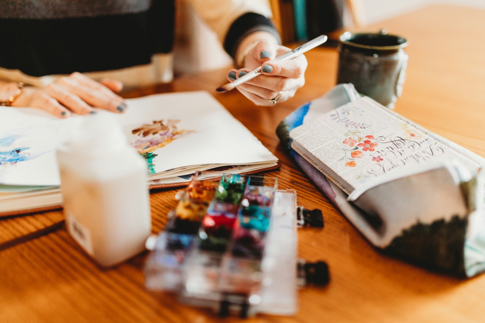 A desk with watercolour paints, a sketchbook and a brush in hand — the materials Paintredo picks for a painting
